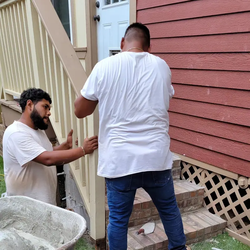 Two crew members working on porch steps beside a red-sided house