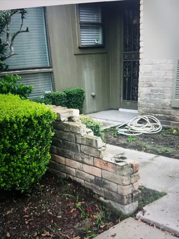 Low brick wall beside a front walk partly collapsed, with loose brick on the ground