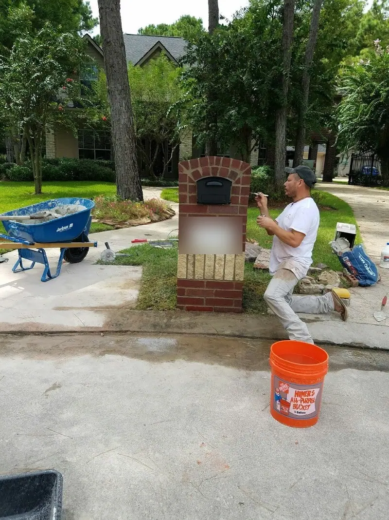 Mason working on a brick mailbox at the curb, with a wheelbarrow and mortar bucket nearby